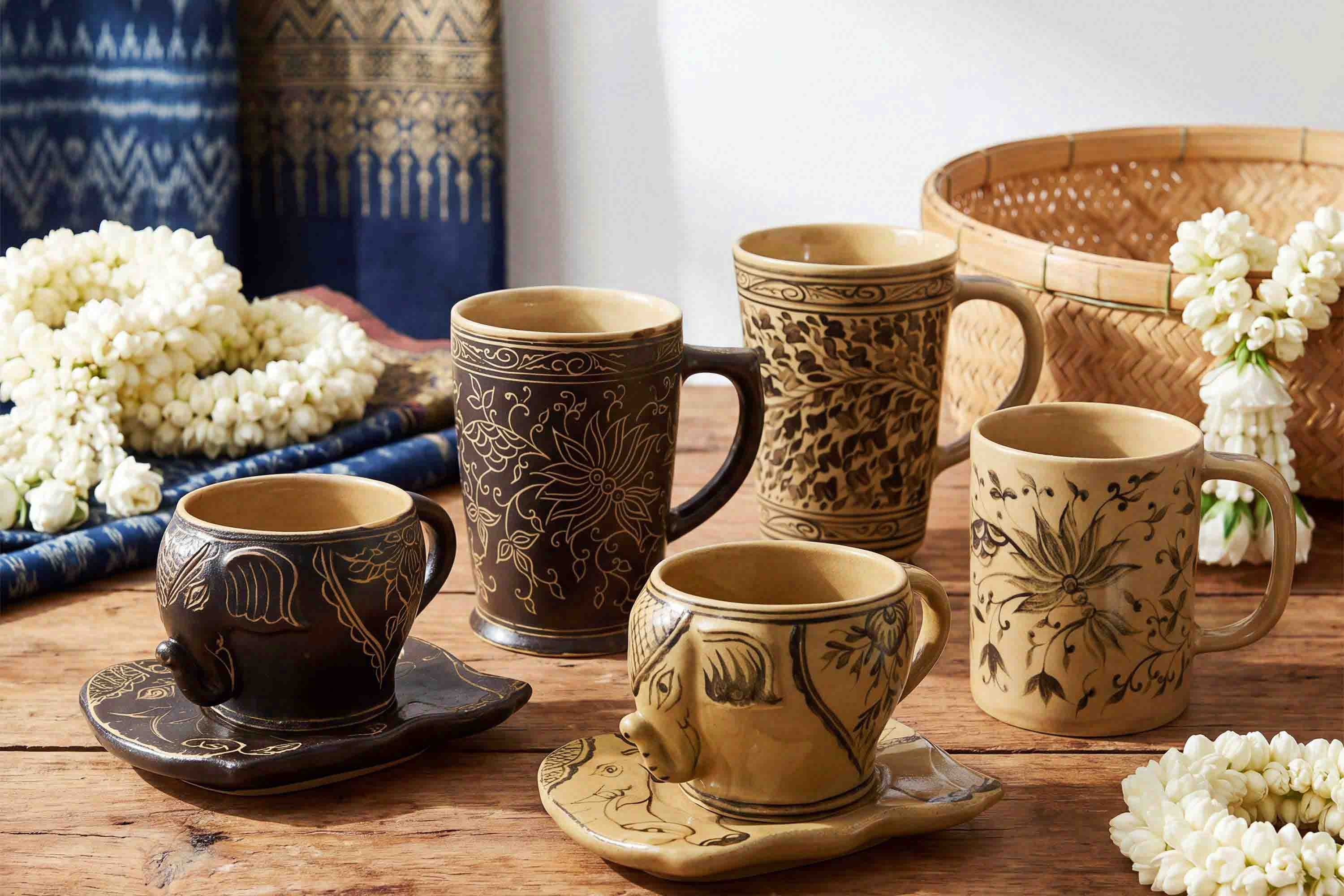 Four decorative ceramic mugs and matching saucers arranged on a rustic wooden table, with white flowers and a woven basket in the background.